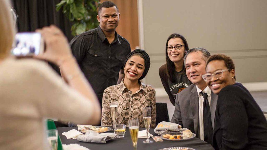 A group of alumni around a table pose for a photo at a Comets Take Flight event. Food and drinks are on the table in front of them.
