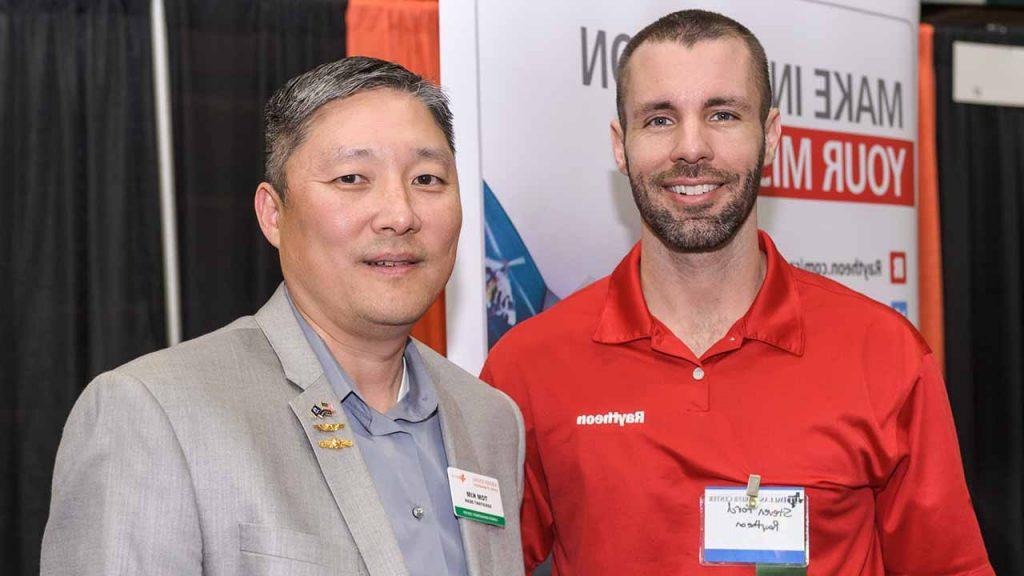 Two people at a career expo on campus. One is representing Raytheon and wearing a branded polo shirt. Both wearing name tags. 