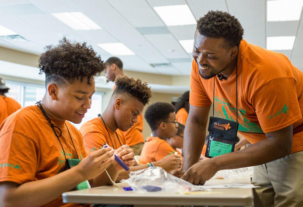 A counselor and several students, all wearing orange t-shirts chat over a table at a camp event. The students are working on a craft with their hands. 